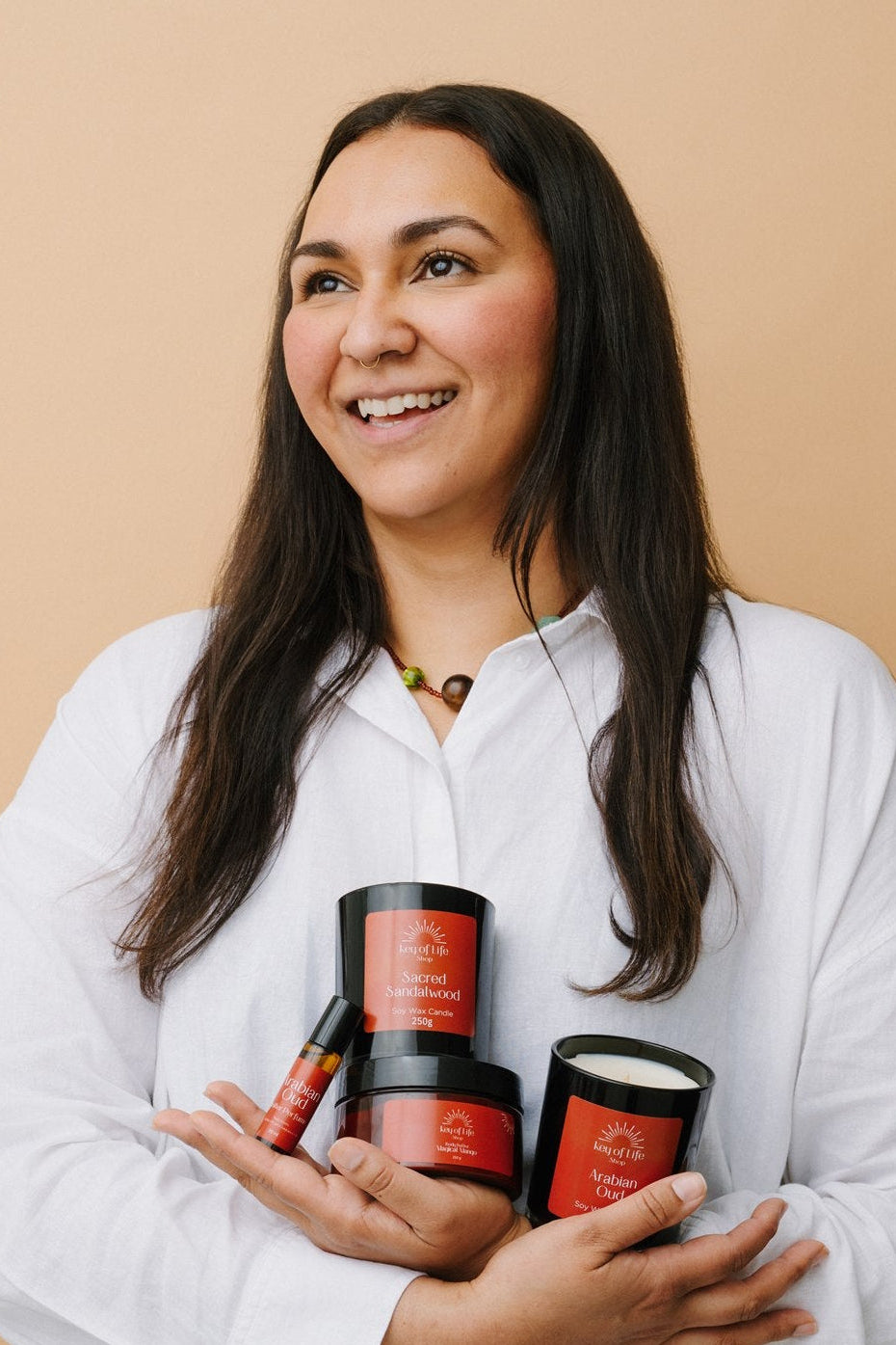 A woman with long dark hair, wearing a white button-down shirt, holds a stack of three red and black cosmetic jars against a solid beige background.